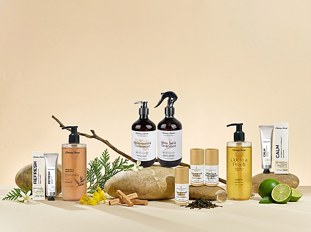 保留原图浅暖米色背景、天然鹅卵石和枯树枝的自然摆台结构，移除原图所有原有产品，替换为参考图中的四组新产品，保持产品大小比例协调，利用原有高低石结构布局让产品错落有致，保留图片上方充足空白空间方便后续添加文字，自然点缀添加雪松枝干、檀木片、新鲜青柠、干茶叶和橙花元素，打造有空气感的高级氛围感香氛系列产品摄影图，调整图片比例为4:3，保持柔和自然光影