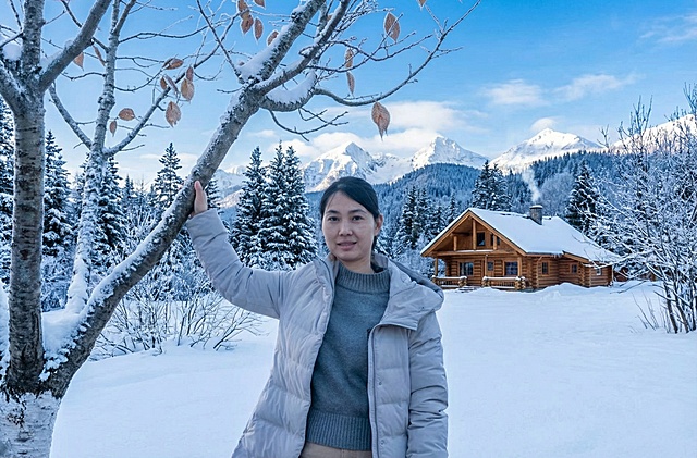 背景中可以看到积雪覆盖的地面、树木、一座小木屋以及远处的山脉，天空呈蓝色并飘着少量云朵，整体色调以蓝白为主，营造出清冷纯净的冬日氛围。