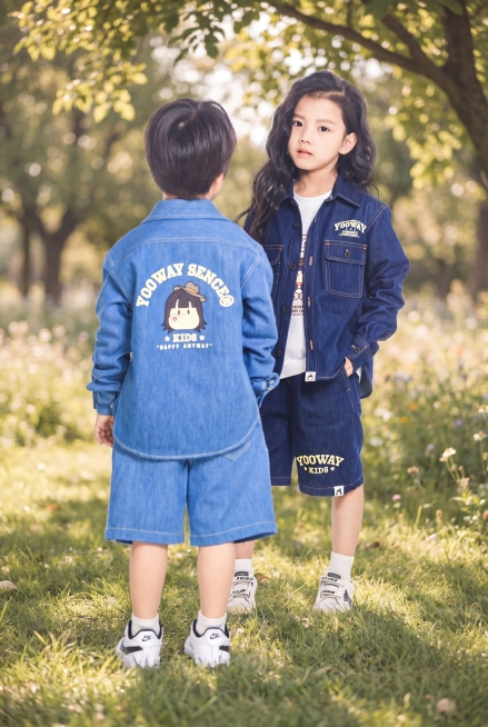 保持图中两个儿童模特和蓝色牛仔童装产品的外观、结构、人物姿态、所有文字和Logo绝对不变，仅替换背景。生成舒适户外自然草坪背景，阳光透过树叶形成斑驳光影，整体氛围清新治愈，符合儿童服饰调性，低对比度柔预览效果