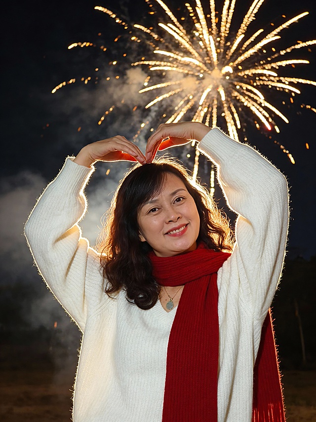 高清8K节日温馨风夜景写真，中年女性，甜美愉悦的节日气质
发型：黑色短发，烟火的逆光打量头发丝
妆容：清透甜美妆（自然通透底妆+淡粉柔雾唇色，面带温柔微笑，神态轻松愉悦）
服饰：白色宽松针织毛衣（面料柔软蓬松，针织肌理清晰），搭配红色长款针织围巾（围巾自然垂落，质感温暖）
身体姿态：站立于户外，身体微微舒展放松，肩颈线条柔和，头部俏皮状稍侧向一边，双手跨过头部做一个比心的形状，姿态随性又带节日的愉悦感
场景：夜晚户外背景，夜空绽放着金色绚烂烟花（带有朦胧烟雾效果），整体氛围热闹温馨
风格：暖调夜景光线打光，高清细腻质感，突出针织面料肌理、烟花的绚烂细节，温馨治愈的节日氛围感