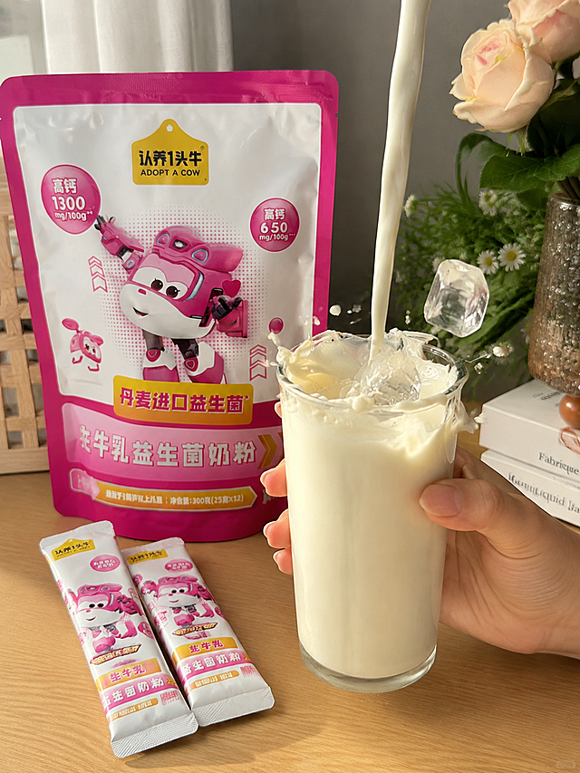 奶液冲调画面，乳白色奶液正注入透明玻璃杯中，冰块落入牛奶中溅起奶花，一只手握着杯子，旁边摆放认养一头牛粉色超级飞侠联名益生菌奶粉袋装和条装产品，居家桌面场景，真实手机拍摄质感，暖光自然光，生活感，3:4竖版构图