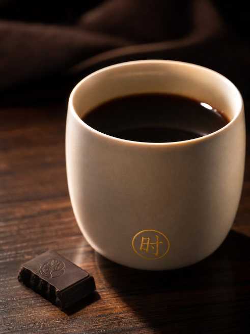 侧光特写，奶米白色厚壁陶瓷杯，侧逆光勾勒出圆润杯身轮廓。杯中咖啡液深褐近黑，液面反射一点高光，放置在深檀色木纹桌面上，杯旁放半块黑巧克力，杯底印有一枚金色"时"字印章，整体氛围浓郁高级预览效果