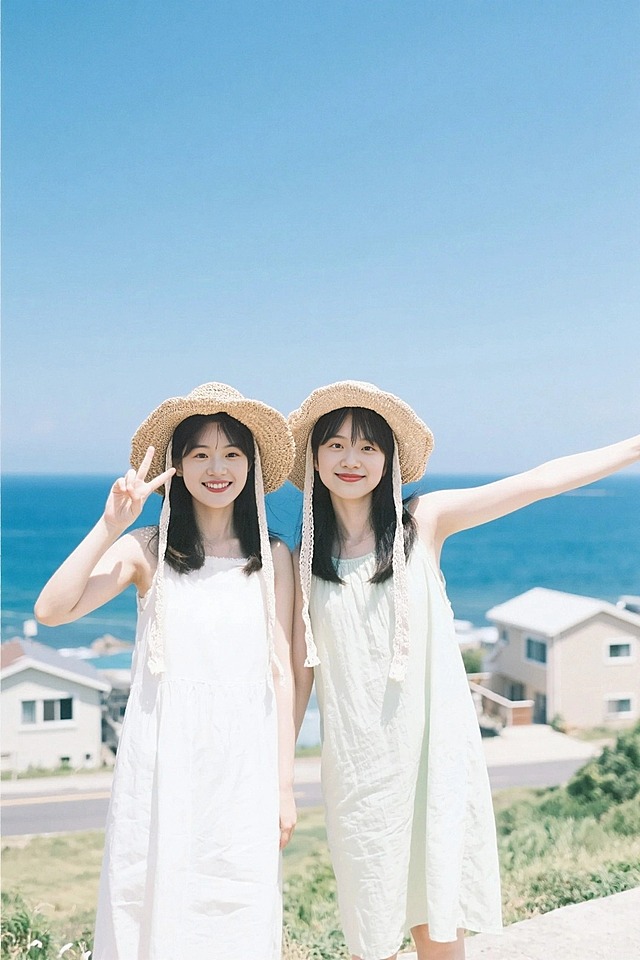 背景济州岛，两位女生戴草帽，穿清新夏装，左边女生比耶，右边女生张开手臂，背景有蓝天、大海、房屋，日系清新色调，胶片质感，远景