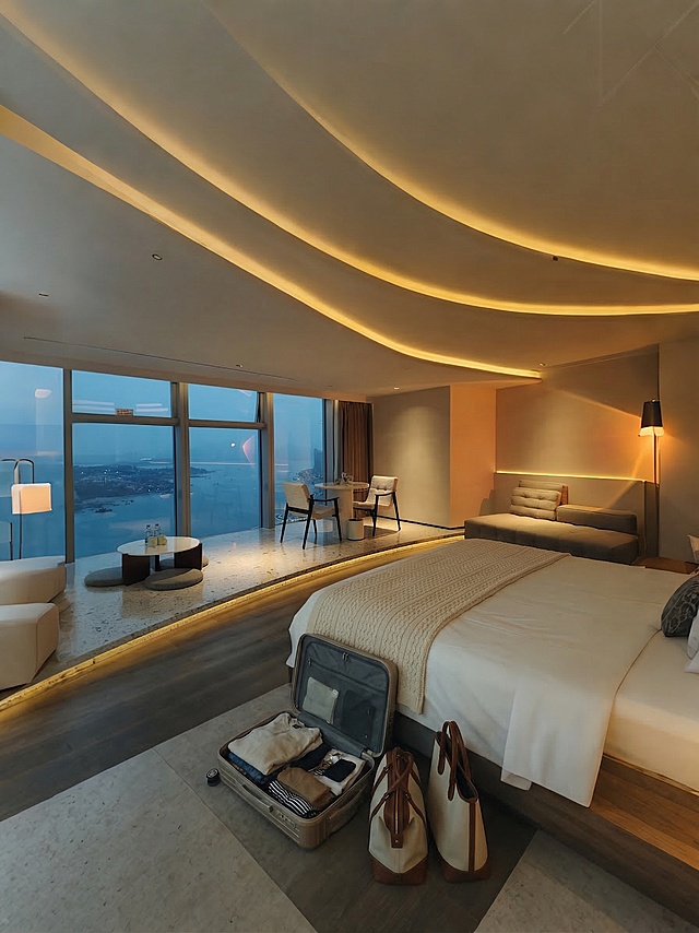 在床上放置一条毯子，在床边地面放置一个打开的行李箱，在行李箱旁边地上放两个手提袋，保持原有现代轻奢海景酒店风格，整体构图光线和氛围不变