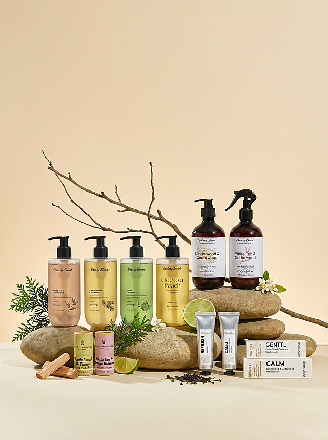 保留原图浅暖米色背景、天然鹅卵石和枯树枝的自然摆台结构，移除原图所有原有产品，替换为参考图中的四组新产品，保持产品大小比例协调，利用原有高低石结构布局让产品错落有致，保留上方充足空白空间方便后续添加文字，自然点缀添加雪松枝干、檀木片、新鲜青柠、干茶叶和橙花元素，打造有空气感的高级氛围感香氛系列产品摄影图，调整图片比例为3:4，保持柔和自然光影