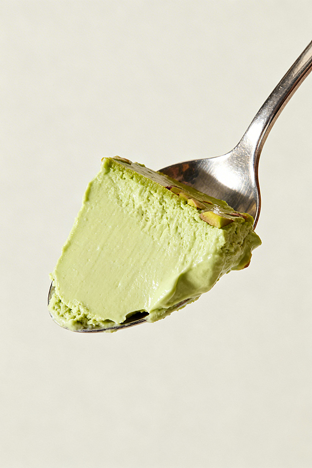 正侧面视角的金属勺子，勺子上盛放着一整块开心果味酸奶酪，凝酪质地，其中一面是平整的，色泽呈现自然的开心果淡绿色，质感细腻浓厚