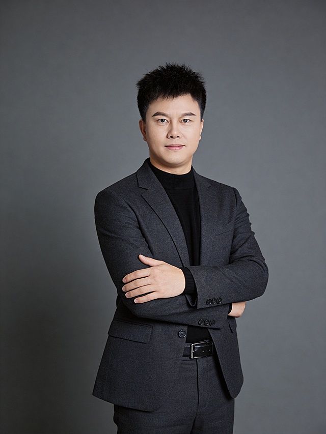 参考图中保留人物脸型、发型、年龄等特征生成职场形象照,标准焦段50mm,f/4光圈(f/2.8-f/4区间),平视视角(镜头与人物视线齐平);人物占画面70%,人物居中(居中构图),位于画面中心,为视觉聚焦点,入境范围头部及以下至臀部下方10cm(半身像);左右留少量背景空间;头部与画面顶部保留5%空间;头部发型完整,头肩比例协调,人物结构准确; 身穿修身精纺羊毛混纺面料的深灰色西装外套(含5%桑蚕丝提升柔光质感),平驳领(商务感正式感),左胸袋无口袋巾(简化高级感),西装下摆自然垂度;内搭黑色中领紧身针织上衣,下装精纺羊毛混纺面料的深灰色高腰直筒西裤(面料同西装),搭配黑色哑光头层牛皮皮带(宽1.5cm),带扣为长方形拉丝黑色金属(1.8cm×2.5cm);着装正式干练;专业稳重;身形匀称,姿态端正,身体挺拔。呈正面抱胸的自然站立姿态:左臂搭于右臂上方,肩颈自然松弛下沉,姿态自信从容;发质微微柔顺，微微蓬松。简约冷调深灰色纯色背景(呈现均匀柔和质感),背景光线均匀柔和,强化色彩对比的高级感。极简干净;画质超高分辨率、细节清晰,现代精致感风格;肤质精细化处理;眼神直视镜头,目光坚定且澄澈,表情自然。嘴角上扬;肤质自然细腻,肤色健康均匀,皮肤状态清爽干净,保留真实皮肤细节｡正面柔光补光,4800K中性光,面部光比1:1.5｡中近景写实摄影,对焦清晰,面部雾面柔光,轻透裸肌感,高清保留皮肤细节｡分辨率4K,去除人物之外的所有元素。严格按照图片内容生成。画面比例3:4