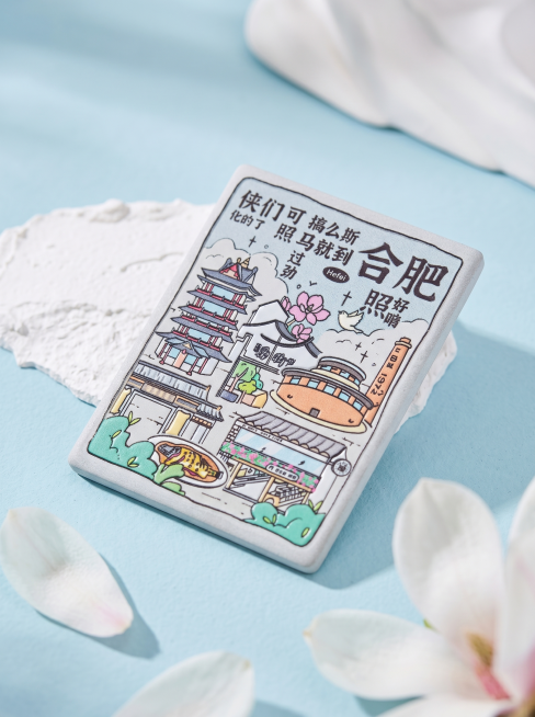 参考图一特写静物构图，清新国风文创产品拍摄，将图2的合肥长方形冰箱贴斜向居中摆放在局部白色石膏上；背景为柔和浅蓝哑光平面，前景点缀少许虚化白色玉兰花花瓣，形成自然前景层次；远景保留局部白色石膏，采用柔预览效果