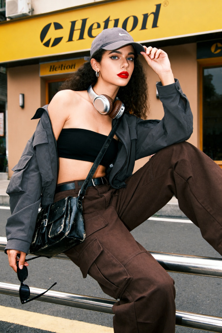一位超高颜值的酷飒叛逆的街头的黄皮法国女生，长相小众，个性十足，一位街头潮酷风少女，戴着灰色棒球帽，颈间挂着复古头戴式耳机，妆容精致冷艳，红唇亮眼。动作是单腿搭在金属栏杆上的随性坐姿，一只手拿着墨镜，预览效果