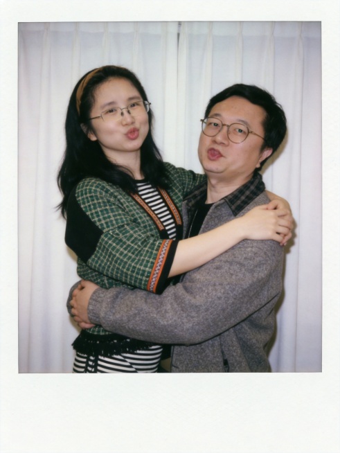 图1人物抱着图2人物，面部细节完全保留与原照片一致，使用参考图中的衣服，背景替换为质感自然的白色窗帘，呈现宝丽来相机拍摄效果，暗室闪光灯均匀柔和打光，画面清晰度一致，两人做搞怪可爱的鬼脸表情，拥抱互动预览效果