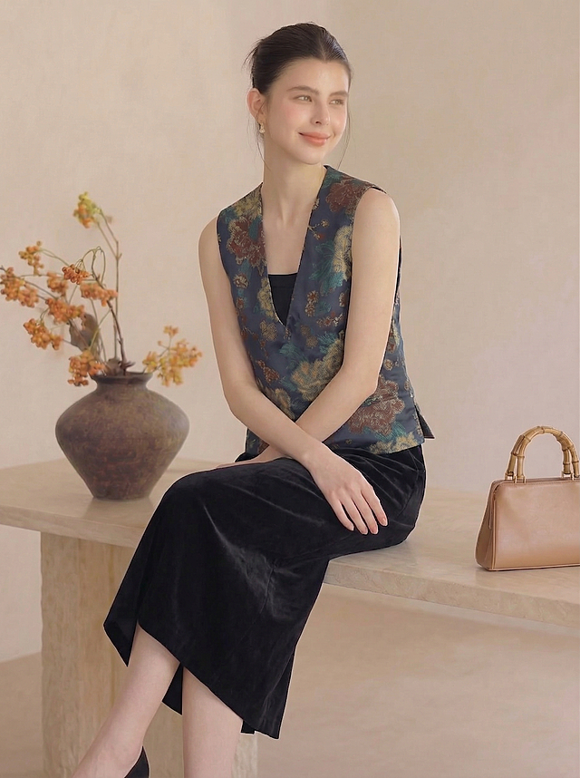 保持人物主体不变，保持深蓝色底彩色花卉提花马甲和黑色丝绒半身裙服饰不变，生成一个自然松弛的全新姿势，保留原米白墙面、陶罐花艺和皮包场景，保持暖调光影和真实质感