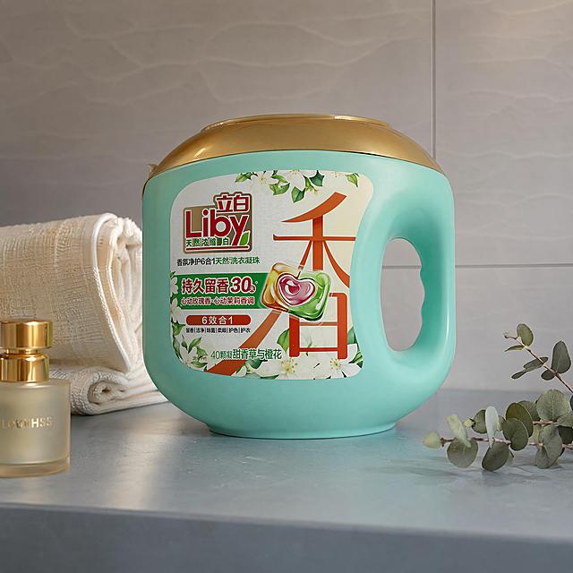 超写实商业产品摄影。
主体：立白洗衣凝珠盒占画面70%，完整不裁切，绝对视觉主导，近景景别；洗衣凝珠盒严格参照上传产品图，完整保留所有可见组件、部件、轮廓、结构、比例、材质、颜色与产品本体上的文字信息（logo/型号/规格/参数等），与上传图完全一致；产品提手、金色顶盖清晰可见，完好无变形。
构图与陈列：微侧15°平视，产品直立放置在浴室哑光岩板洗手台边缘，平稳接触无悬浮。
风格：现代轻奢风。
场景概念：轻奢浴室洗漱区，暖光室内灯下。
承载与背景：①承载面：雾霾蓝色哑光岩板台面；②背景：浅灰色鱼肚纹瓷砖墙面。
陪衬物：①画面后方，一块亚麻纹理擦手巾，米白色亚麻材质；②台面左前方，一只金色迷你香薰瓶，磨砂玻璃搭配金色瓶盖；③台面右角，一小株尤加利绿植，灰绿色叶片。
配色：主60%雾霾蓝+浅灰，辅25%米白+灰绿，点缀15%金色；颜色为雾霾蓝、浅灰、米白、灰绿、金色；材质为岩板、瓷砖、亚麻、玻璃、金属、绿植。
光影：主光来自斜前45°暖光室内灯，辅光为墙面柔光反射，瓶身塑料质感细腻，有自然柔和高光。
参数：4K 写实光追。严格锁定宽高比为 1:1。