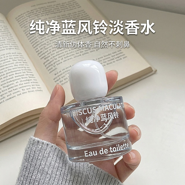 严格还原参考图中的香水瓶产品，保留所有包装设计、颜色、LOGO位置、文字内容、图案元素细节，产品为迷你便携款（约高6-8cm，宽4-5cm），主视觉为身穿浅灰色针织毛衣的手手持香水瓶特写，背景是ins风浅米色书桌搭配翻开的英文书籍，整体极简清冷风格，画面干净通透，光影柔和，8K高清，细节丰富。添加文字：主标题"纯净蓝风铃 淡香水"加粗放大居中，副标题"清新伪体香 自然不刺鼻"小字点缀在主标题下方，文字使用简约无衬线白色字体，排版为精致杂志风。