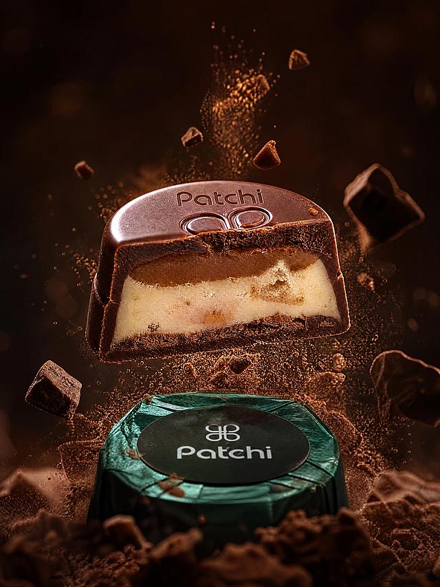 高端商业摄影产品海报，主体是Patchi巧克力，完整保留参考图中墨绿色锡纸包装圆形巧克力的外观和切开的多层夹心剖面层次，伦勃朗光式电影级明暗对比，低角度仰拍强透视，微距镜头感，巧克力碎屑与可可粉飘散的动态画面张力，深棕黑色极简抽象背景配微弱光斑，奢华精致高级的奢侈品广告摄影风格，Octane渲染，4K超高清，巧克力丝滑光泽、夹心酥脆层与锡纸金属肌理极致真实，大师级低饱和高级配色，居中对称完美构图，艺术感诗意氛围感，获奖级视觉冲击力，产品绝对主体突出，浅景深虚化，超写实高细节