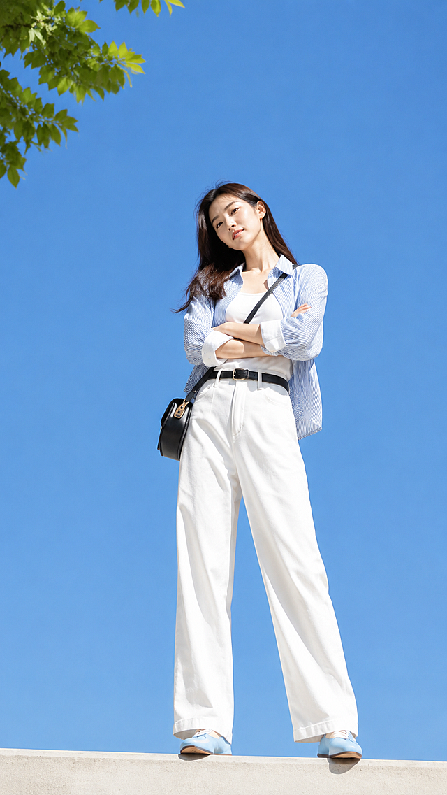 竖版全身照，低角度仰拍，1位年轻东亚女生，棕褐色柔顺中长发，清淡素颜妆容，双腿自然分开站立，双臂交叉环抱胸前，身体微侧，头部歪向一侧，慵懒看向镜头，站在水泥平台；穿搭：蓝白细竖条纹宽松翻领衬衫，袖口外翻露出白边，内搭白色修身背心，白色高腰阔腿长裤，黑色针扣腰带，黑色斜挎软皮包，蓝白拼色休闲板鞋；背景纯净湛蓝色晴空，画面左上角少量绿叶，晴天户外自然光，通透日光，富士胶片写实，8K高清，衣物纹理清晰，皮肤细腻，清新韩系穿搭写真，画面明亮干净