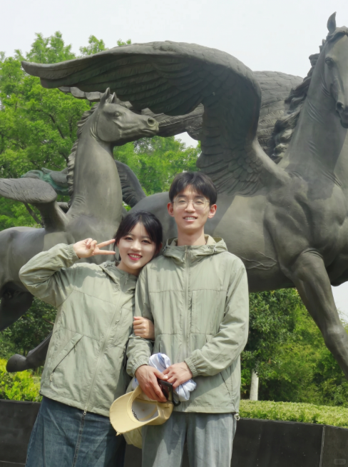 将图2的灰绿色连帽外套分别穿到图1飞马雕像前这对情侣身上，男女都换上这件灰绿色外套，替换掉原来的衣服，过渡自然平整，保持原来的雕像公园背景和合影姿势，图片比例3比4预览效果