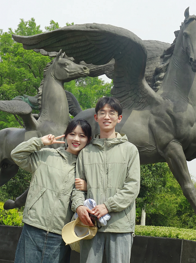 将图2的灰绿色连帽外套分别穿到图1飞马雕像前这对情侣身上，男女都换上这件灰绿色外套，替换掉原来的衣服，过渡自然平整，保持原来的雕像公园背景和合影姿势，图片比例3比4