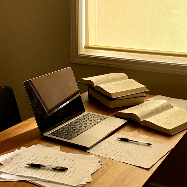 真实摄影风格，图书馆靠窗的学习书桌桌面，更加松弛自然的凌乱桌面，摆放着打开的笔记本电脑，摊开的厚书籍，散落着写满笔记的纸张、笔，木质桌面纹理清晰，窗外自然光柔和漫射进来，真实的生活质感，无人出镜，现代预览效果