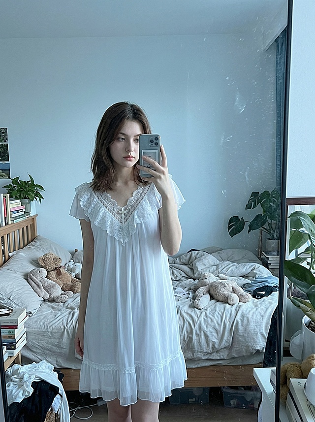 基于参考图片中的白色睡裙，生成一张中国年轻女生穿着这件睡裙对镜拍的图片，保留衣服的材质和精致细节，尤其保持花边的精致轻薄；场景为杂而有序的居家背景，床上放着几个玩偶；风格为朴素的日常图片，不需要专业的景深，色调清透，偏冷色调；镜子上带有一些灰尘，女生五官端正，身材纤细，留着日常中长发