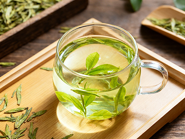 透明玻璃杯中冲泡好的明前龙井茶叶，汤色清澈嫩绿，几片鲜嫩茶叶舒展在水中，放在素雅的木质茶盘上，周围点缀少量干茶和茶荷，清新雅致，自然光下的茶文化摄影，简约干净的构图