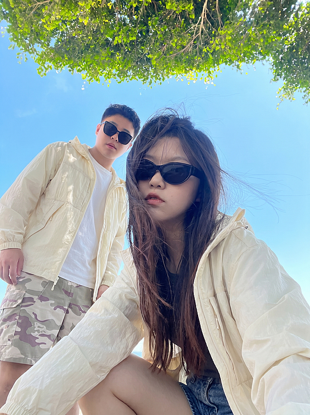 将图2的米白色连帽外套分别穿到图1这对男女身上，两人都换上这件米白色外套，替换掉原来的衣服，过渡自然平整，保持原来的蓝天树荫背景和人物姿势，图片比例3比4