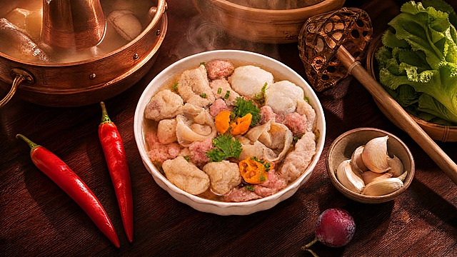 什锦汤碗严格参照上传产品图，完整保留碗的造型、汤品、鱼丸、肉片、肉燕、豆泡、黄辣椒、香菜、葱花、红辣椒、小果子、蒜碟等所有可见组件、轮廓形状、结构、比例、材质质感、颜色与原图完全一致，自然融入新场景光影。
潮汕牛肉火锅店场景，深红木色调，浓郁鲜香氛围。
背景为深红木质餐桌，左上角可见铜制牛骨汤锅与砂煲，右侧搭配竹制漏勺与新鲜生菜，蒸汽升腾。
暖调暖光侧照，产品受光面泛起红棕色、暗部映射深红木色光晕，与环境物理融合。
保持原图视角和构图不变，产品位置居中。
4K分辨率，严格锁定宽高比与原图一致。
