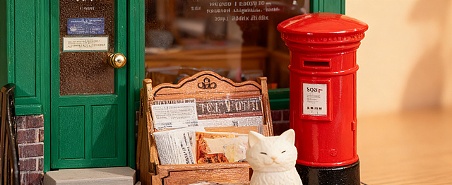 近距离特写展示邮局小屋的绿色门、白色小猫、红色邮筒的组合区域，清晰呈现门的磨砂质感、金属门把手光泽、小猫的可爱造型纹理、邮筒的亮面材质，三者同时入镜，保留暖光桌面环境，光影柔和自然，尺寸为宽1464px高600px。