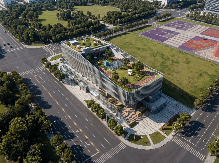 基于图1生成摄影级鸟瞰效果图，保持图1原有建筑角度和建筑形体不变，不添加额外建筑。参考图2的建筑造型和色彩，制作高透光玻璃幕墙搭配铝板造型外墙。保留周边城市环境和铺装广场，维持原有画面角度，整体色调为预览效果