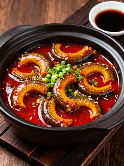 经典淮扬菜红烧马鞍桥，黄鳝段切成马鞍形状，色泽红亮诱人，裹着浓稠鲜亮的红烧酱汁，软嫩的鳝肉纹理清晰，表面撒上翠绿的葱花和少许白芝麻提香，盛放在哑光黑砂锅里，摆放在深色木质餐垫上，旁边放一小碟香醋蘸料，预览效果