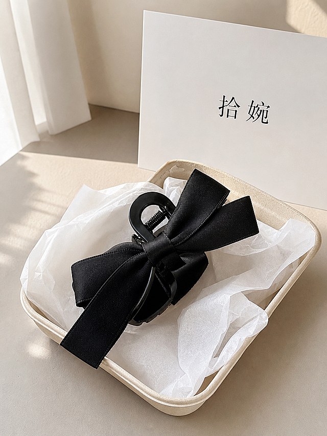 俯拍平铺静物摄影，赫本风黑色蝴蝶结抓夹女大号高级感盘发网红发夹后脑勺鲨鱼夹头饰，放置在米黄色可降解纸浆盒内，盒内衬白色薄棉纸；盒子后方摆放白色纸质卡片，卡片印有黑色文字:“拾婉”；干净浅米色桌面，柔和窗边自然光，韩式简约饰品静物，低饱和莫兰迪色调，高清电商实物实拍，浅景深，面料刺绣纹理清晰细腻，高清，画面简洁高级