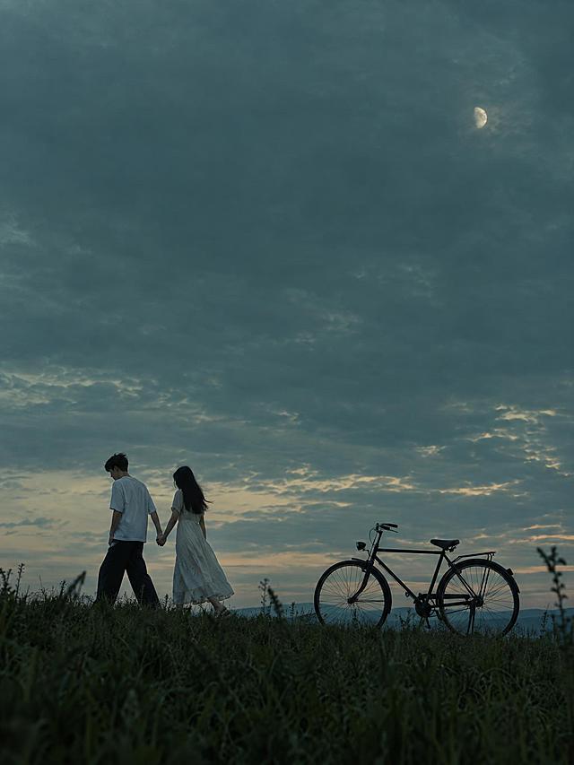 竖版3:4电影感写实静帧，低饱和青绿色暮色。广阔阴天占画面上方三分之二，右上方一轮朦胧小月亮；低机位草坡上，一对二十多岁的东亚年轻恋人牵着手向左侧缓缓走去，男生穿宽松白色短袖和深色长裤，女生长发、浅色连衣裙。人物位于画面下方偏左，右侧停着一辆复古黑色自行车，车轮完整可见，前景是深色草丛剪影。安静、克制、略带忧郁的浪漫氛围，柔和暮光，自然真实摄影质感，上方保留干净天空留白。画面中不要任何文字、不要标题、不要字幕、不要Logo、不要水印。超高清2K画质，细节丰富。