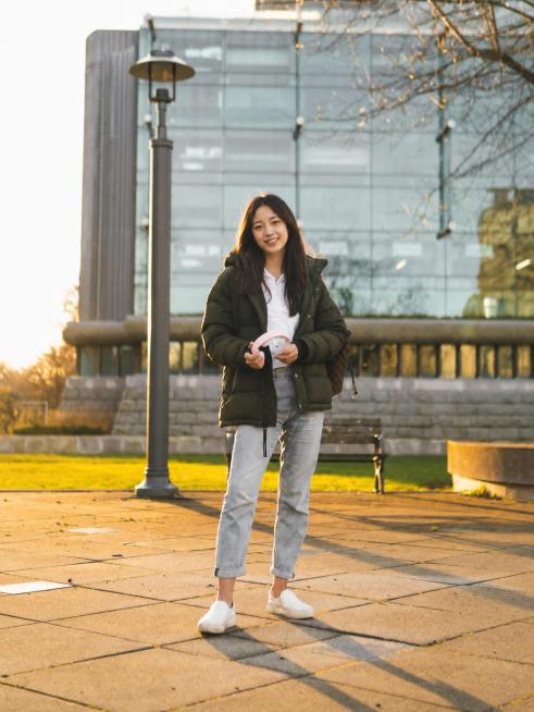 把图片中的女生换成真实自然的中国女生，保持原有姿势、穿着（深绿色羽绒服+白色内搭+浅灰色牛仔裤+白色鞋子）、背景和整体光线氛围不变，效果真实自然预览效果