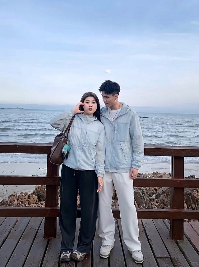 将图2的浅天蓝色连帽外套分别穿到图1海边栈道这对情侣身上，男女都穿上这件浅蓝外套，替换掉原来的上衣，过渡自然平整，保持原来的大海栈道背景和姿势，图片比例3比4