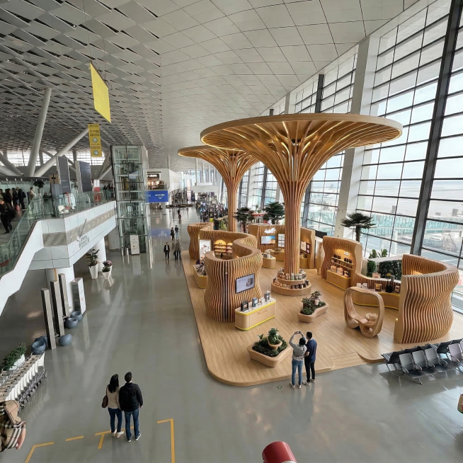 修改这张机场商业空间设计图，将两个柱子做得更矮、更小，整体颜色调整为原木色与米白色相互融合，在整个设计区域下方增加一块浅色木质抬高地板，保持原布局和空间通透感，不遮挡右侧落地窗，写实摄影风格预览效果