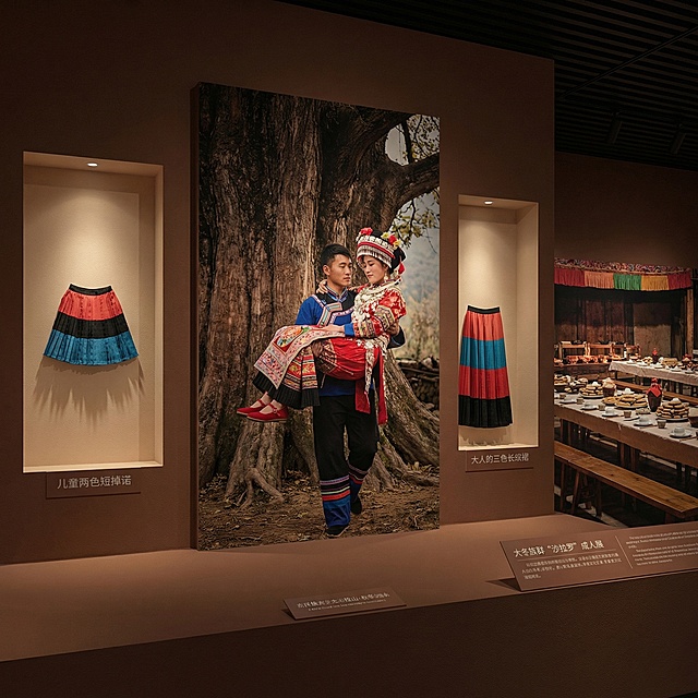 彝族沙拉洛成人礼民俗展陈场景，背景为秋冬季节的大凉山高山林间，画面中心呈现表哥背负着盛装少女走到高大古树下举行象征性树婚的核心仪式场景，少女神态羞涩又充满期待。场景龛内左侧展示二色短款童年百褶裙，右侧展示三色拖地成年长裙，形成实物对照。背景宴席区域的长桌上摆满苦荞粑粑、苦荞酒等传统饮食，凸显苦荞作为仪式核心见证的文化角色。整体氛围庄重喜庆，充满民族文化厚重感，光影温暖柔和，符合民俗展陈的叙事需求。
