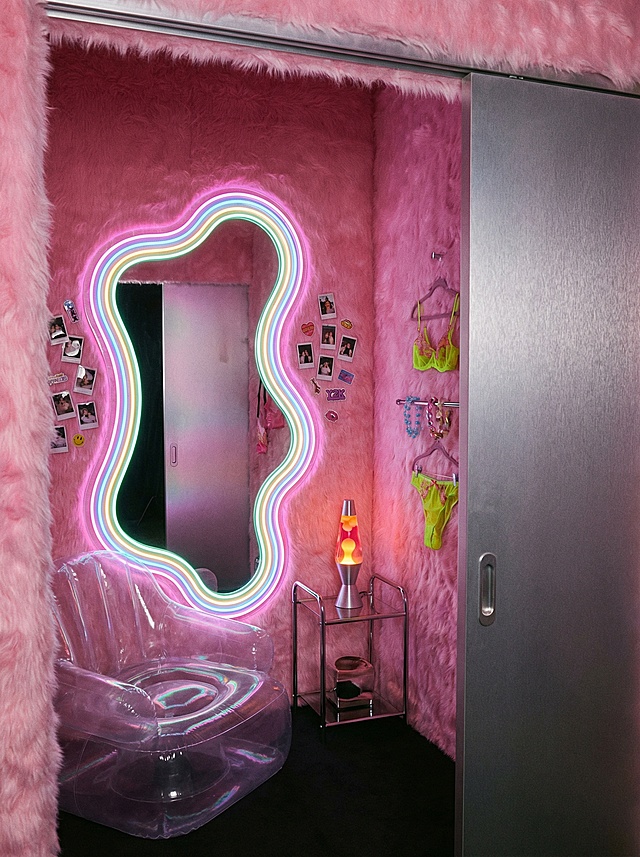 A luxurious and playful lingerie store fitting room, Y2K-inspired. The walls are covered in plush, pink faux fur. The mirror is not a standard rectangle, but a large, amoeba-shaped, full-length mirror with a glowing neon frame. Inside, a small, transparent, inflatable chair sits in the corner. The lighting is soft and flattering, provided by a retro-style lava lamp on a small chrome shelf. The door is a sleek, sliding panel of brushed aluminum. The vibe is intimate, fun, and makes you want to take a selfie.