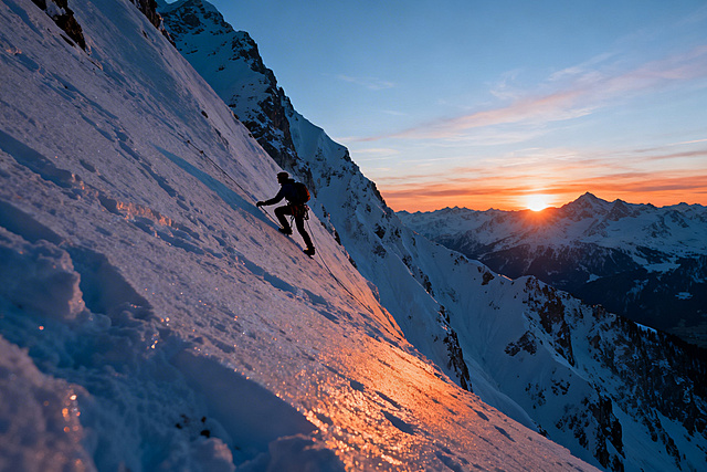 攀岩雪山的人物侧身远景照，人物在陡峭雪壁上攀爬，远处雪山后太阳微微升起，霞光晕染天际
