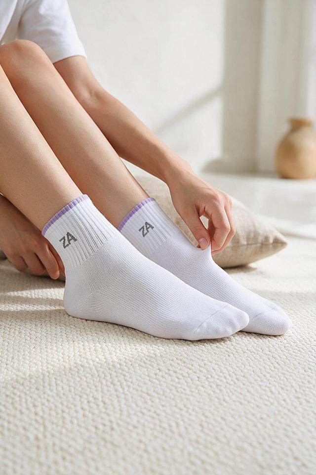[商品] 使用【图1】【图2】【图3】【图4】【图5】中的白色+浅紫罗纹袜口针织短筒袜；100% 继承原图的针织面料、纱线纹理、织物颗粒、原有刺绣印花图案、褶皱走向与明暗光影，袜子质感完全复刻原图；保留罗纹袜口撞色边、袜头袜跟加固结构与白色棉纱本色，袜身网眼透气织法不变。商品处理：自然理顺袜身与袜口织物，去除杂乱压痕与松垮褶皱，增强纱线与针织颗粒细腻质感，不改变材质、颜色与结构，禁止重新生成袜子材质。
[画面] 目标：韩系 ins 风模特场景图，展示袜子真实穿着全貌与整体氛围。模特自然坐姿，双脚穿着白袜向前舒展并轻轻交叠，一只手自然搭在脚踝、另一只手轻捏袜身整理，动作自然、符合人体结构。袜子位于画面视觉中心，占画面约 65%。中景略俯视，浅景深。前景米白色哑光棉麻布面，中景模特足部与袜身，后景柔和虚化的米白墙面与低饱和莫兰迪道具（亚麻靠垫、浅驼色陶器）。
[视觉] 套系策略：风格=韩系 ins 静物生活摄影；主色=米白；背景延展=基于米白做深浅层级与棉麻织物、纸张质感；强调色=浅紫（袜口罗纹）与浅驼；光影=柔和侧方自然光。低饱和莫兰迪色调，极简留白，干净高级，浅景深，细腻面料纹理，柔和阴影，无反光，画面无杂物。
[文字] 画面中不出现任何文字、标题、卖点、水印、Logo 或排版元素。
[规格] 图片比例 2:3 竖版，2K 高清商品摄影，8K 级细节。负向：人脸、畸形手脚、卡通、文字水印、袜子变形、色彩失真、杂乱杂物、蓝灰冷调背景、材质被重新生成。