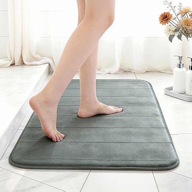 图上的珊瑚绒记忆棉地毯是我的产品，请使用我的产品帮我生成亚马逊商品图，构图高级，富有张力，电影质感，1600:1600比例，超高清：
核心元素：浅灰色记忆棉浴室地垫，一双白皙光脚轻踩地垫，一只脚落地、一只脚抬起；背景有白色大理石瓷砖、白色浴帘，旁置白色洗护瓶、浅灰托盘与干花装饰。
场景：明亮现代浴室，干净整洁，暖光氛围，凸显舒适居家感。
构图：中景俯拍，地垫居画面中心，脚部为视觉焦点，背景元素简洁不抢镜，构图平衡。
色彩：主色调浅灰 + 白色，点缀干花暖色调，色调柔和明亮，质感细腻。
风格：高清写实电商产品图，光影柔和，突出地垫柔软纹理与防滑质感。
文字：高清写实电商场景图，细节清晰，无文字，纯产品展示，聚焦视觉卖点，排版简洁干净。