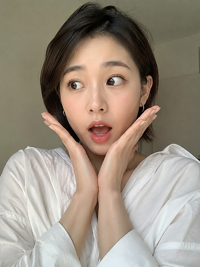 居家近景自拍，半身像，气质短发女生，简约造型耳环，白色宽松上衣；神态夸张惊讶，眼睛向侧面看，双手抬起摊在脸颊两侧，简约干净浅色室内背景，柔和暖调自然光，高清写实博主人像