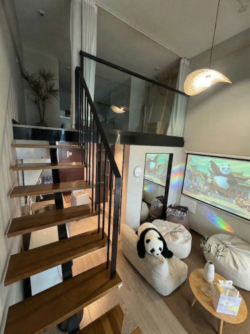 基于现有loft复式客厅图片，在米色沙发上放置一个熊猫造型抱枕，在木质小圆桌上放置一个简约陶瓷花瓶。保持原图整体空间结构不变，保留木质楼梯、玻璃护栏、编织吊灯、功夫熊猫投影、现有豆袋沙发等原有家具结构预览效果