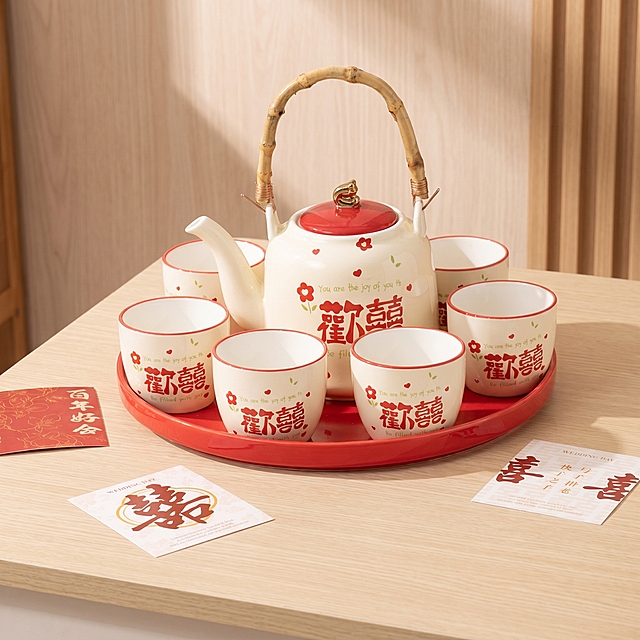 [主图 1] 欢囍陶瓷茶具五杯款家居场景主图，去除所有多余文字；
[氛围风格] 温馨舒适家居治愈风，轻奢高档质感；
[画质] 商业摄影质感、光线柔和通透、产品清晰真实、画面干净高级，超高清画质满足2.8M大小需求；
[元素] 欢囍陶瓷茶具整套（1茶壶5茶杯带礼盒）居中饱满陈列在浅米白色桌面，红色茶盘衬托，茶具保持米白底配红色欢囍文字的外观，物理结构、材质质感、颜色与输入产品图绝对一致，杜绝任何变形和色差，精细化处理。保留周边小喜字卡片装饰，背景是浅木色墙面，整体氛围感温馨治愈。移除画面中所有多余的宣传文字，仅保留茶具本身的欢囍文字。商品主体占比严格控制在 70% 左右，画面满屏无边框；
[光影] 明亮柔和的侧窗自然光，均匀通透，陶瓷釉面呈现温润高光，木色桌面漫反射出温暖层次感，光比舒适，暗部细节清晰，无过度曝光；
[配色] 主色调为米白暖木色，点缀红色文字与茶盘，整体温馨干净；
[视角构图] 平视取景，产品完整居中展示，景深适中，画面层次清晰；
[比例分辨率] 1:1 比例，2k 分辨率，文字清晰可辨，超高清；
[负向提示词] 变形，水印，多余文字，额外宣传文字，文字错乱，产品图模糊，背景杂乱，夸大违禁词，遮挡主体
