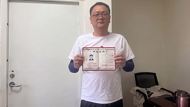 请保持参考图1中人物的面部、发型、眼镜、白色T恤、黑色裤子、左臂深色袖套、站姿、表情、背景（白色门、办公椅、桌子、暖水壶、墙面）、光影、构图、景别完全不变；仅将人物手中的毕业证书面积缩小约50%，同时双手距离同步变窄、双手向中间收拢以自然持握缩小后的毕业证，手指抓握姿势自然协调；毕业证书仍保持正面朝向镜头、水平展开，1:1还原其红色封皮、文字、印章、黑白照片、细节纹理、污渍质感等所有细节，尺寸符合真实高中毕业证比例，与双手大小比例协调自然。

超写实高清画质，细节层次丰富，色彩还原真实，整体质感自然真实；毕业证书与手部接触处有自然接触阴影与压痕，边界清晰、层次分明、接触点自然贴合。