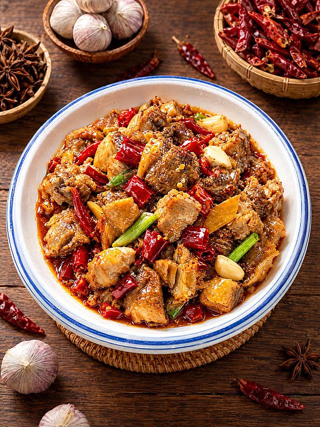 基于参考图中的红烧鸡块菜品，生成60度俯拍角度的美食摄影图，保持菜品色泽红亮、辣椒葱段蒜瓣配料丰富，蓝边白瓷盘装盛，木质桌面背景，背景有独蒜、干辣椒、八角等香料，暖色调专业美食摄影，高清细节，诱人食欲