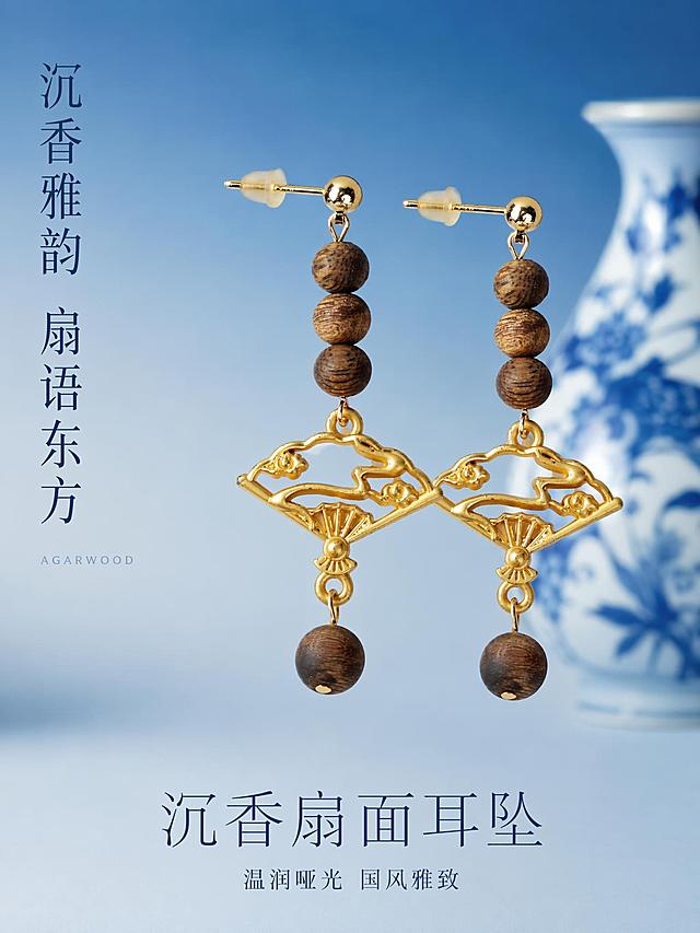 将图一中的沉香耳坠完整保留，套用图二的清新渐变蓝背景+青花瓷瓶东方雅致国风摄影风格。耳坠珠子数量、珠径、天然木纹、油线分布、深浅色泽、串型结构、金色镂空扇面祥云吊坠、金色耳针必须与原图完全一致，保持温润哑光沉香原生质感，拒绝塑料光泽和过度提亮。竖版3:4构图。画面构图：一对沉香耳坠并排悬挂展示，左侧耳坠稍靠前，右侧耳坠略靠后形成层次感，位于画面中上部分，背景为浅蓝到深蓝的渐变哑光背景，右后方有青花瓷瓶虚化作为装饰，底部留有充足负空间，整体氛围清新雅致东方国风感。光影：柔和侧光从左上方照射，形成自然柔和的阴影，沉香珠子表面呈现温润哑光质感，油线纹理清晰可见，金色扇面吊坠温润金属光泽，无强烈反光。配色：主色调渐变蓝、青瓷白、沉香原木色、金色暖调，整体色调清新雅致有国风韵味。商品主体占画面约45%，居中偏上位置，画面满屏无边框。左侧负空间竖排两行大字"沉香雅韵 扇语东方"，纤细宋体，深蓝色，字号大而雅致；下方极小英文装饰"AGARWOOD"，纤细衬线字体，浅蓝色。底部负空间加文字，主标题"沉香扇面耳坠"大号纤细宋体，深蓝色；副标题"温润哑光 国风雅致"小号同色系，上下排列居中。超高清商业摄影质感，细节锐利，景深适中，背景虚化柔和。