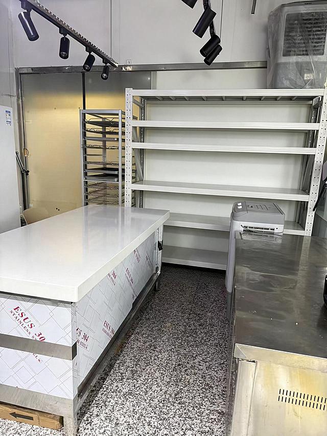 去除后方白色多层货架上的所有物品（包括纸箱、塑料盒、包装袋、瓶罐、篮子等所有摆放在货架各层上的物品），仅保留空的白色货架主体结构，货架的金属框架和层板完整保留。严格保持房间内其他所有主体、细节、结构、颜色、光线、透视角度完全不变，不得添加或修改任何其他元素。去除物品后的区域用周围真实的墙面和货架结构自然填充，光影和材质与原图一致，过渡自然无痕。最后整体提升图片清晰度，画质自然真实，保持原照片的真实感。