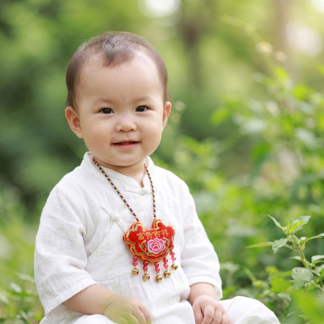 端午节日氛围，户外绿意朦胧背景，一岁穿白色衣服的精致可爱女宝宝，胸前挂着一个图片参考中的红色刺绣富贵吉祥长命锁香包，香包带有金色铃铛装饰，整体画面温馨治愈，1:1构图预览效果