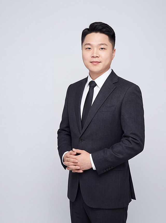 参考图中人物的脸型、发型、五官特征保持不变，生成年轻干净的90后职场精英形象照。标准焦段50mm，f/4光圈，平视微侧视角，镜头与人物视线齐平。人物占画面70%，人物略偏右构图，位于黄金分割点，半身照，左侧留更多背景空间，头部与画面顶部保留5%空间，头肩比例协调，人物结构准确。身穿深炭灰色高级挺阔西装外套，平驳领设计，面料为高支羊毛混纺，质感硬挺有型，哑光高级质感，剪裁利落修身，肩线挺拔；内搭纯白色高支棉衬衫，系黑色哑光窄领带，简洁干练；下装深炭灰色同面料西裤；着装简洁高级，年轻精英气质，身形匀称挺拔。呈侧身微转站姿，身体微侧面向镜头，双手自然交叠于身前，肢体放松自信。发型干净利落，精神清爽。脸部紧致瘦脸效果明显，下颌线清晰锐利，脸部线条利落紧致，脸型更显小巧立体，肤色白皙透亮均匀，完全去黄去暗沉，皮肤干净清透，呈现年轻健康的白皙肤色。纯净冷调浅灰背景，均匀柔和，极简干净，背景光线均匀明亮。画质超高分辨率、细节清晰，现代年轻高级感风格。肤质精细化处理，眼神明亮有神，直视镜头，表情自然带淡淡自信微笑。皮肤白皙细腻，通透有光泽，零黄气零暗沉，哑光清透肤质，保留真实皮肤纹理但更显年轻紧致。杰作，最佳画质，超高细节，写实摄影，中近景人像，皮肤质感清透白皙，明亮柔光，正面均匀补光，画面干净明亮高级，高清皮肤细节。面部光比1:1.5，5200K冷白光，脸部紧致提升效果明显，下颌线清晰，脸型更立体小巧V型，肤质清透白皙，面部柔光，轻透裸妆感，真实毛孔纹理但更细腻。去除所有配饰。
