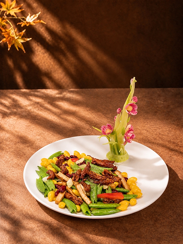 将第一张图中的银杏蜜豆炒鸭舌（白色圆盘装的银杏、蜜豆、鸭舌、豆干、红椒等食材，以及盘内的花卉摆盘装饰）完整替换到第二张图的暖棕秋日背景场景中，替换掉第二张图中原有的白贝芋仔煲菜品、黑色砂锅和木砧板。保留第二张图的全部背景元素：暖棕色墙面背景、左上角的黄色枫叶枝叶、枫叶光影投射效果、暖棕色桌面、柔和侧逆光与光影氛围。白色圆盘直接放置在暖棕色桌面上，不要木质垫板/砧板，位置与视角自然协调，光影与背景侧逆光方向一致，菜品上有自然的光影投射效果，融合自然。不要添加热气蒸汽效果。画面中不要任何文字、菜名或装饰元素，只有背景和菜品主体。竖图3:4比例，写实商业美食摄影质感，食材色泽鲜亮诱人，光影柔和自然，整体秋日暖调氛围统一。