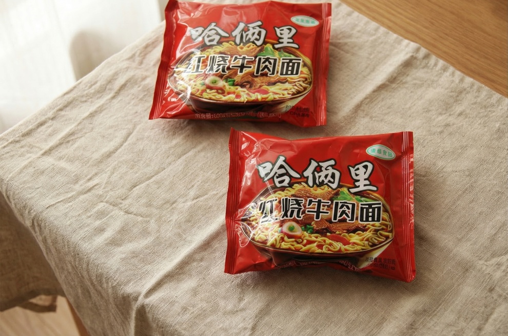 将两包哈俩里红烧牛肉面自然融入铺有棉麻桌布的木质桌面场景中。要求：1. 保持包装袋上的所有文字和Logo原样，保持主体物理结构不变；2. 光影匹配：使用全屋漫射柔光，模拟柔和窗光，生成符合物理规律的淡预览效果